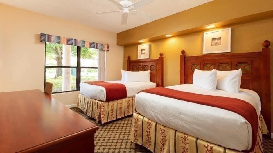 A bright guest room is furnished with two large beds, each with crisp white linens and decorative red accents. A wooden desk is positioned against the wall. Natural light flows through a large window, framed by patterned curtains, offering views of the exterior landscaping.