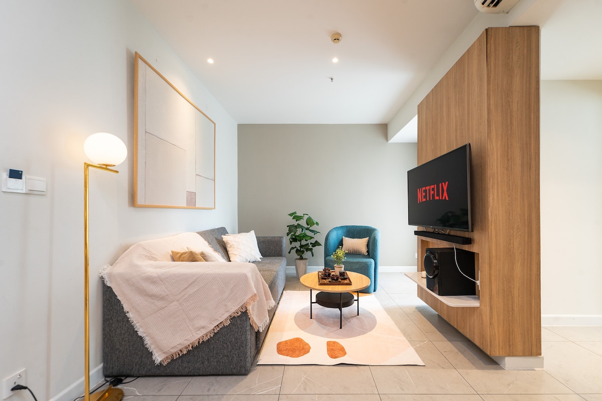 The living room features a spacious sofa draped with a light-colored throw, accompanied by a stylish chair. A wooden TV unit holds a screen displaying Netflix. A round coffee table with decorative items sits at the center, and a plant adds a touch of greenery.