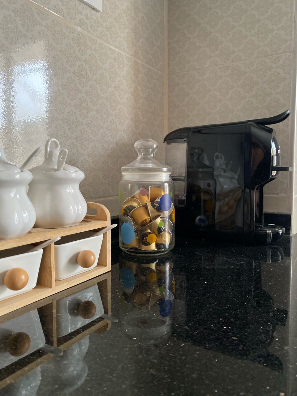 A sleek black coffee machine is positioned beside a glass jar filled with colorful coffee capsules. White ceramic containers with rounded lids sit on a light wooden tray, complementing the reflective black countertop. Subtle floral patterns adorn the light wallpaper, enhancing the kitchen's inviting appearance.