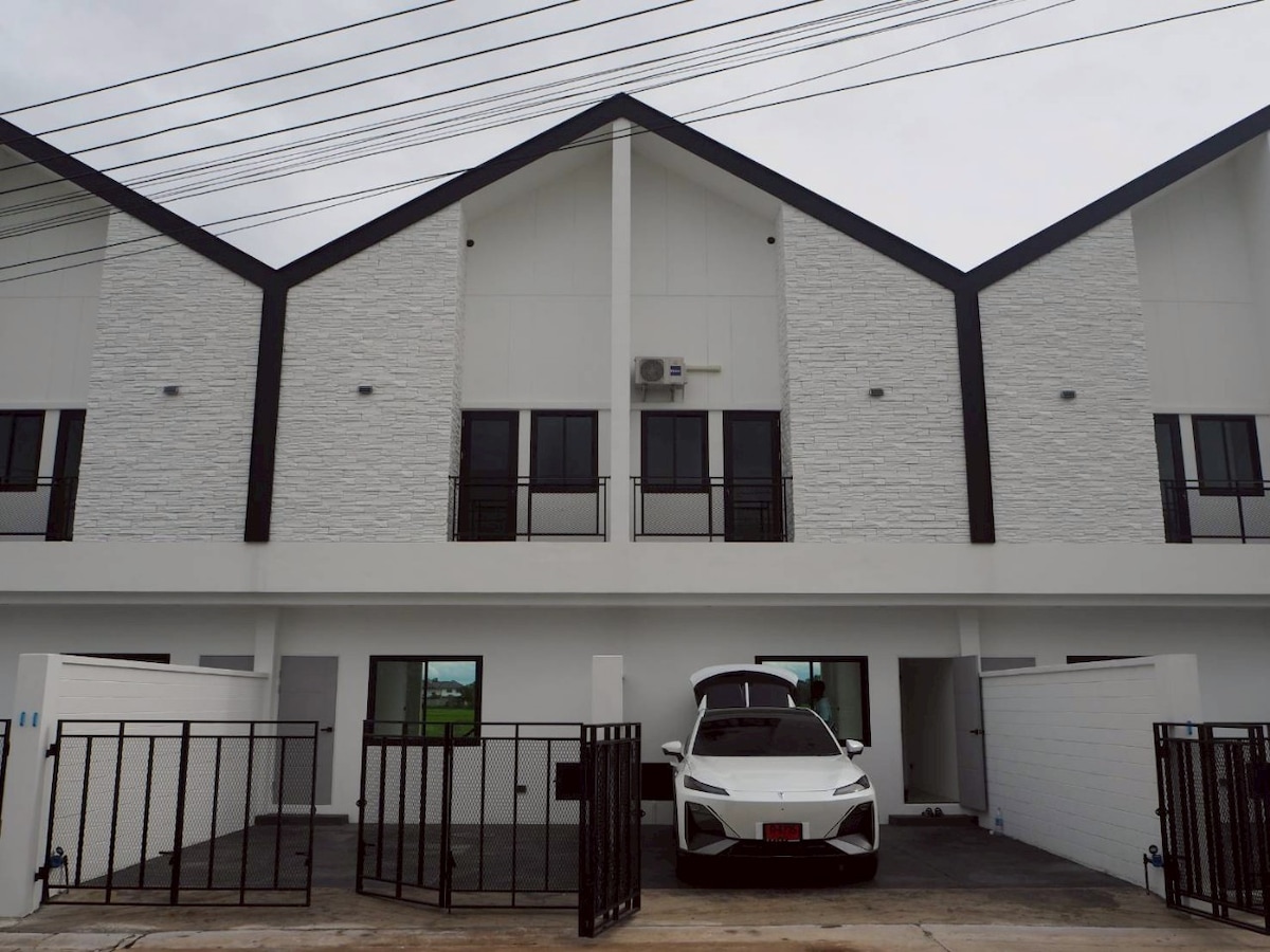 The front view of a two-story townhome showcases a modern design with a light-colored facade. A car is parked in the driveway, which is enclosed by a gated fence. Large windows are visible on both levels, allowing natural light into the home.