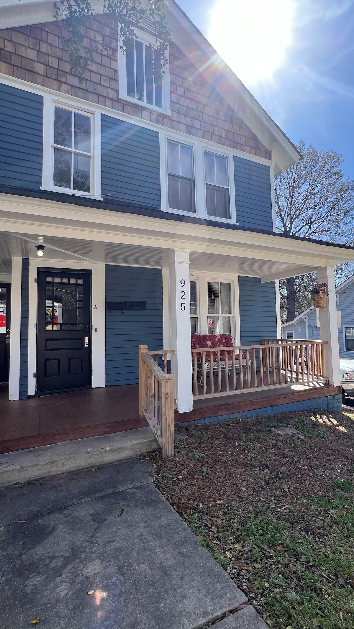A welcoming front porch features a wooden railing and two stylish chairs set against a background of blue and brown siding. The entrance door is black, complemented by a prominent house number. Natural light filters through the trees, creating a bright atmosphere.