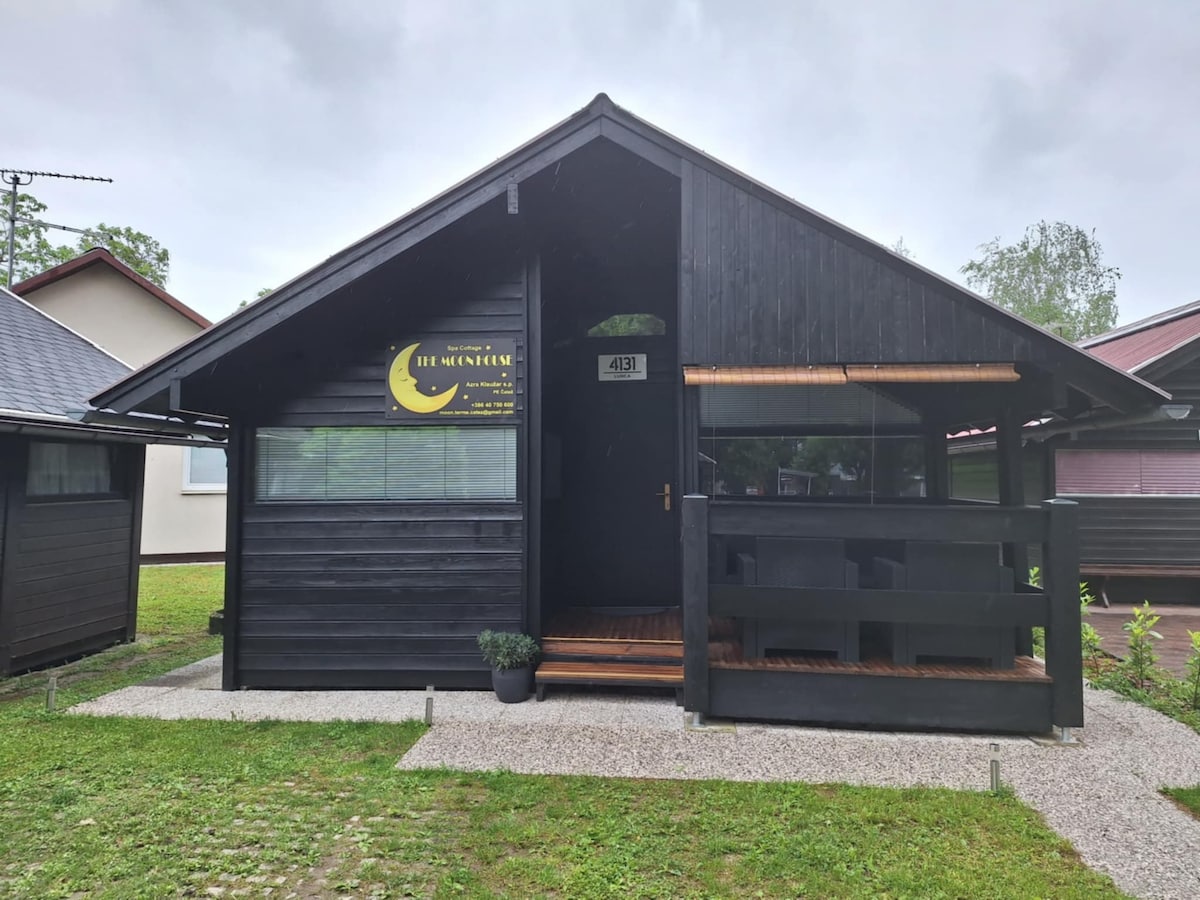 The exterior of a black wooden cottage is showcased, featuring a sloped roof and large windows. A covered porch area is visible, adorned with a simple railing. Lush green grass surrounds the home, contributing to a serene natural setting.