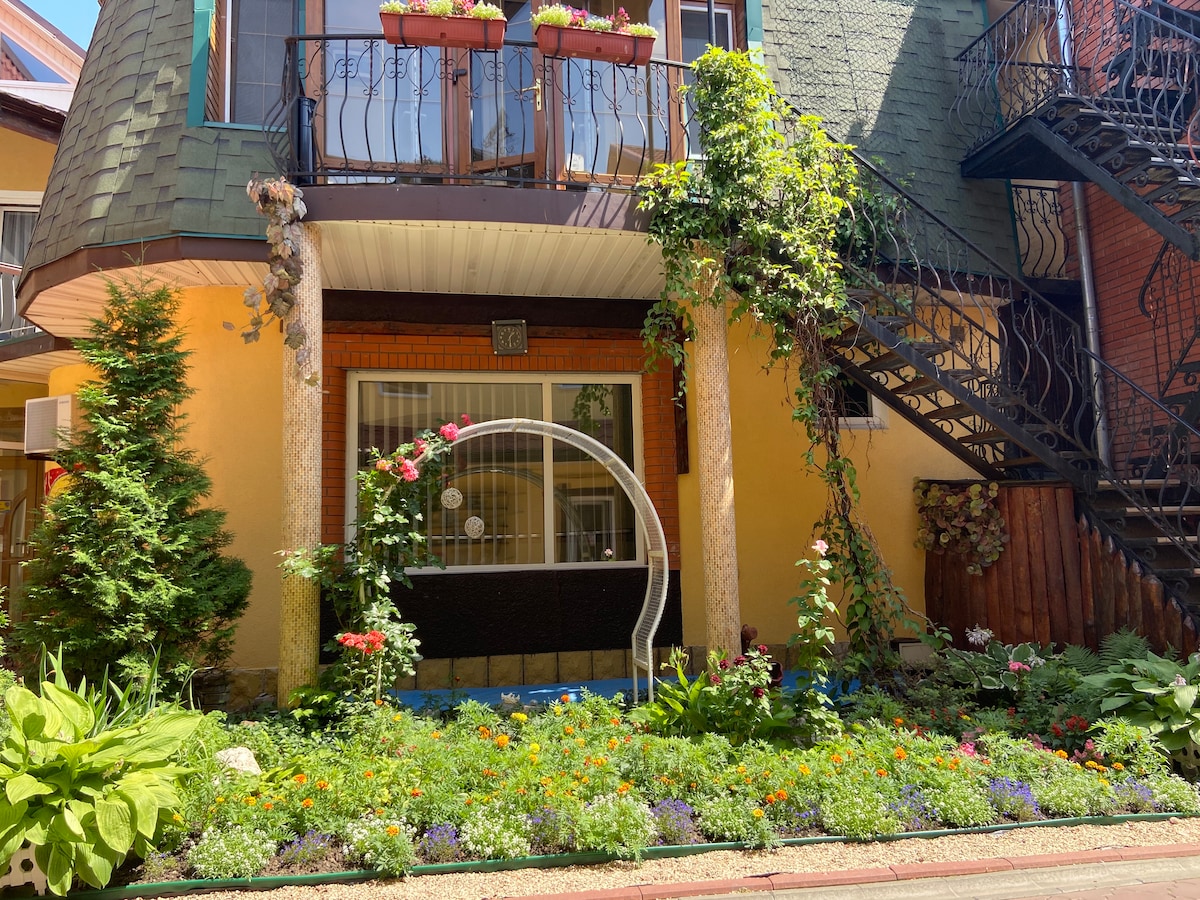 The exterior of a charming hotel is presented, featuring a well-maintained garden filled with colorful flowers. A decorative arch is positioned in front of the entrance, surrounded by lush greenery. A prominent staircase is visible, leading to upper-level accommodations.