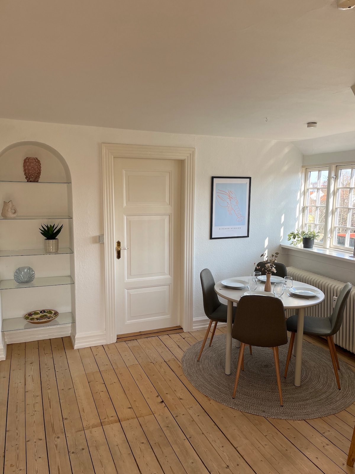 A dining area is presented with a round table set for four, accompanied by modern chairs. Natural light filters through the large window, highlighting a decorative shelf featuring plants and tasteful accents. A door leads to another space, complemented by wooden flooring.