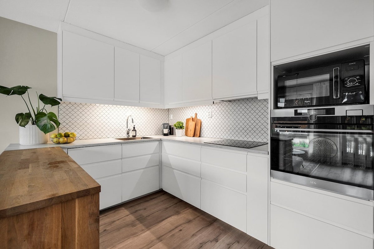 A modern kitchen features sleek white cabinetry and a geometric tile backsplash. A wooden countertop provides workspace, while a small plant adds a touch of greenery. An oven and coffee maker are integrated into the cabinetry, emphasizing the kitchen's functional design.