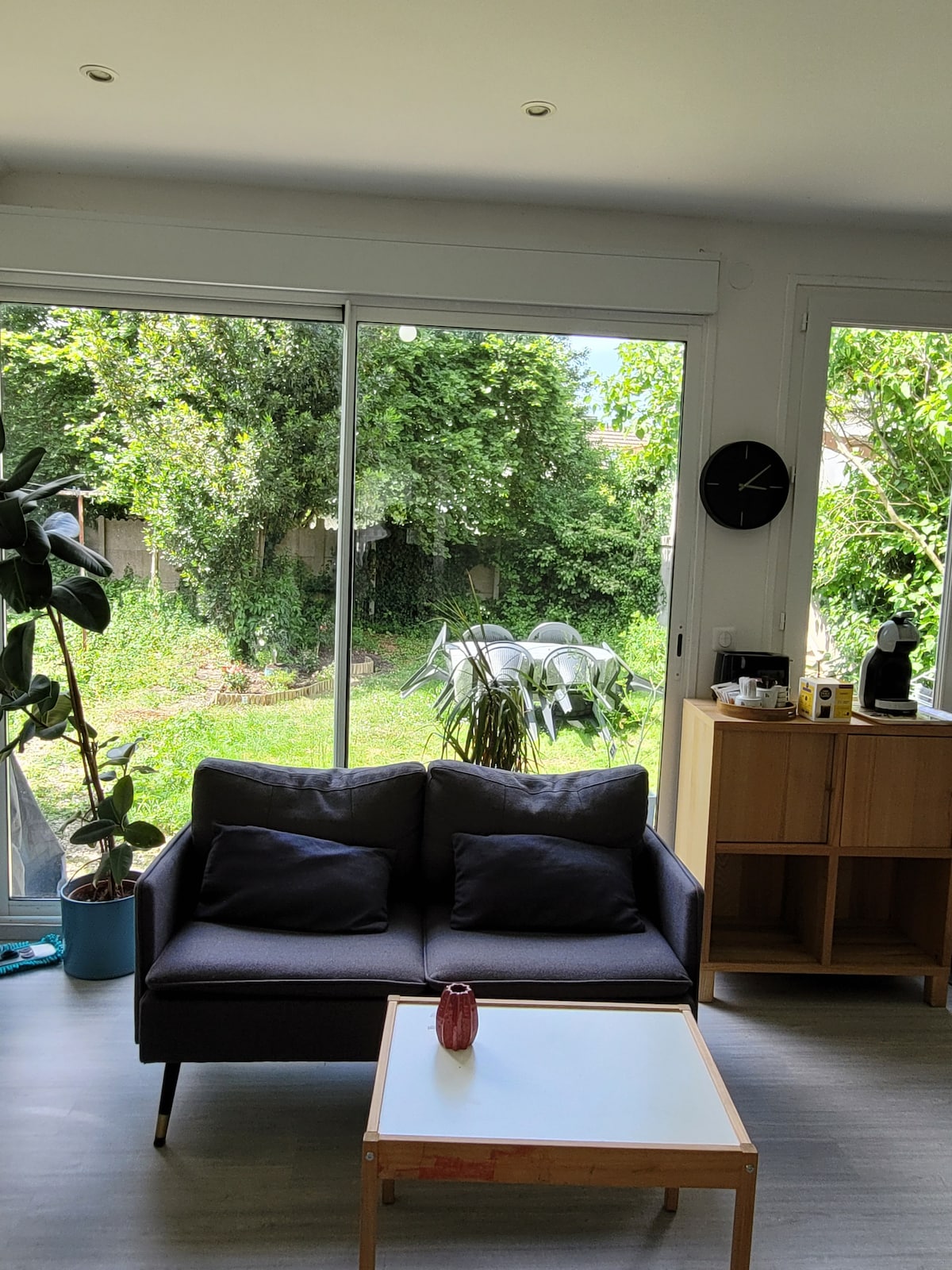 A light-filled living area is presented, featuring a dark sofa paired with a small coffee table. Large glass doors offer a clear view of the lush garden outside, which is enhanced by abundant greenery. A circular clock is mounted on the wall, adding a practical touch.