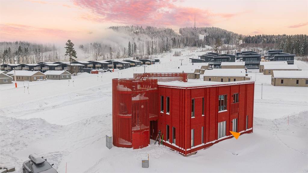 An aerial view captures a modern red building surrounded by snow-covered grounds. Rolling hills rise in the background, partially obscured by mist. Nearby structures are also visible, offering a glimpse of the winter landscape.