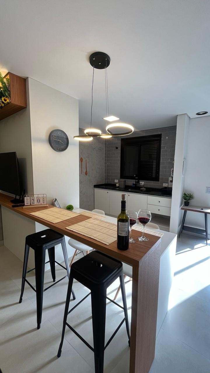 A modern kitchen area features a sleek bar with three high stools. A minimalist dining setup is displayed with two glasses of red wine and a bottle. Soft lighting is provided by overhead fixtures, and the stylish cabinetry is accented by a light and dark color palette.