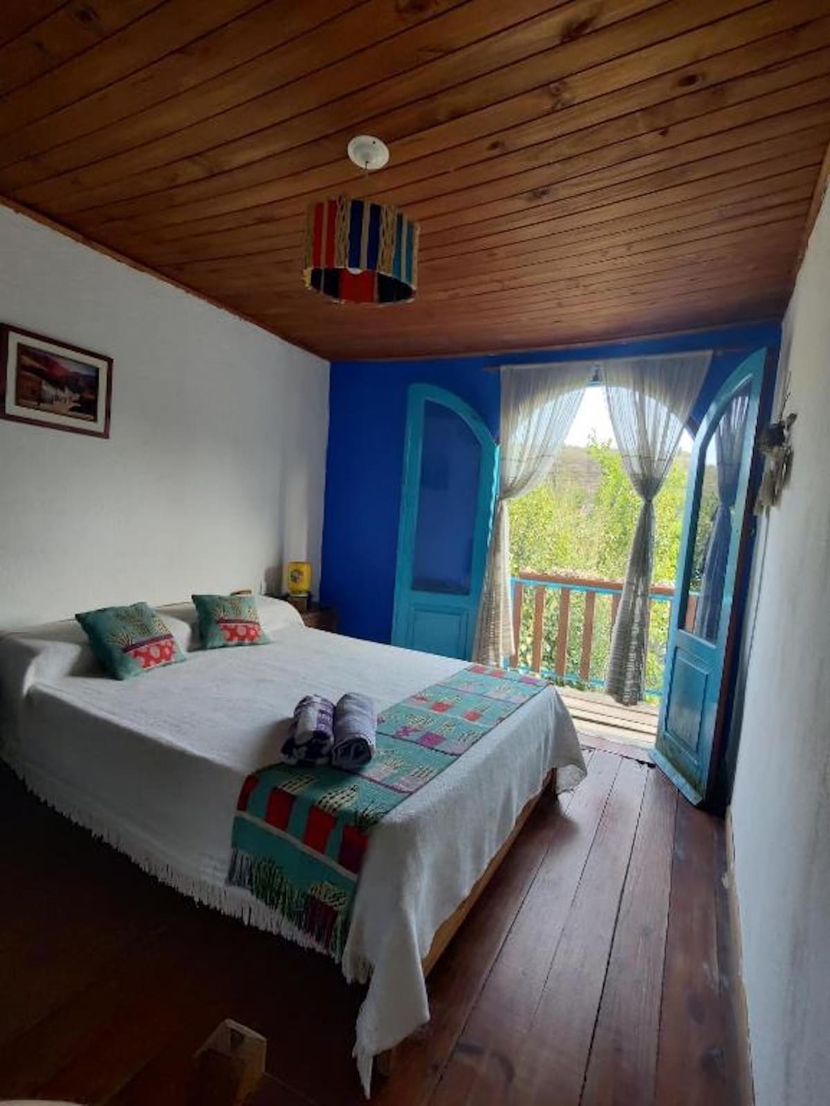 A bright bedroom features a spacious bed with a decorative blanket. Two colorful cushions add a vibrant touch. Large blue doors open to a balcony, with sheer curtains allowing soft light to enter. Natural wood flooring complements the warm wooden ceiling.