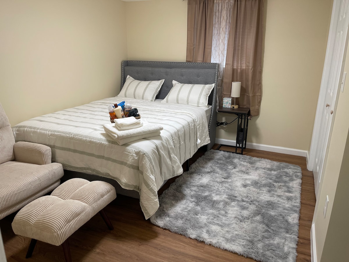 A spacious bedroom features a large bed with white linens and striped gray accents. A cozy armchair and ottoman accompany a soft gray rug on the floor. Natural light filters through a window covered with light curtains, and a small nightstand holds a lamp.