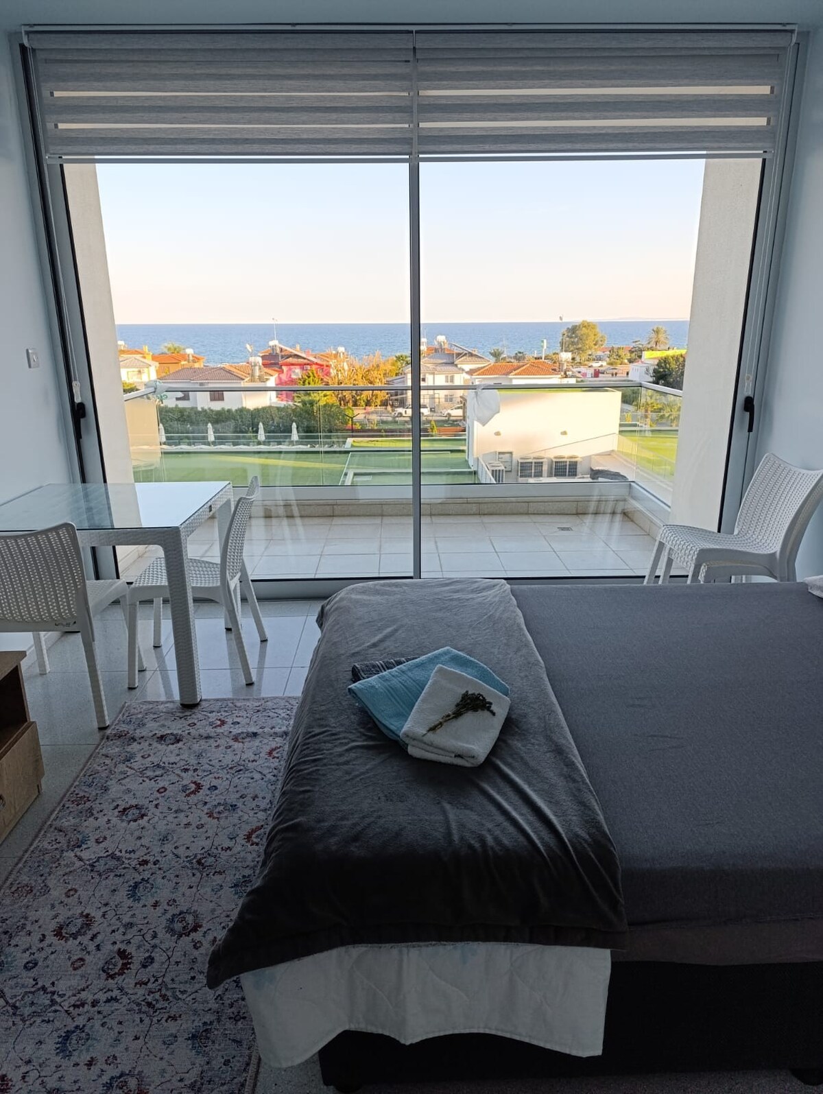 A spacious sleeping area features a large window providing a sea view. A cozy bed is accompanied by a soft blanket and a small decorative item. A dining table with two chairs sits nearby, allowing for a comfortable space to enjoy meals.