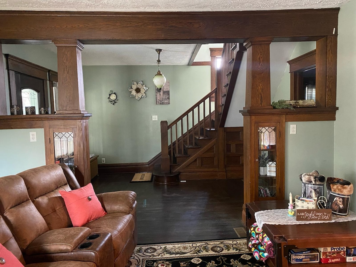 The living area features polished wood trim and old wood floors. A cozy seating arrangement includes a brown leather couch, placed near a decorative table. A staircase with wooden banisters leads to the upper floor, while framed wall decor adds character to the green walls.
