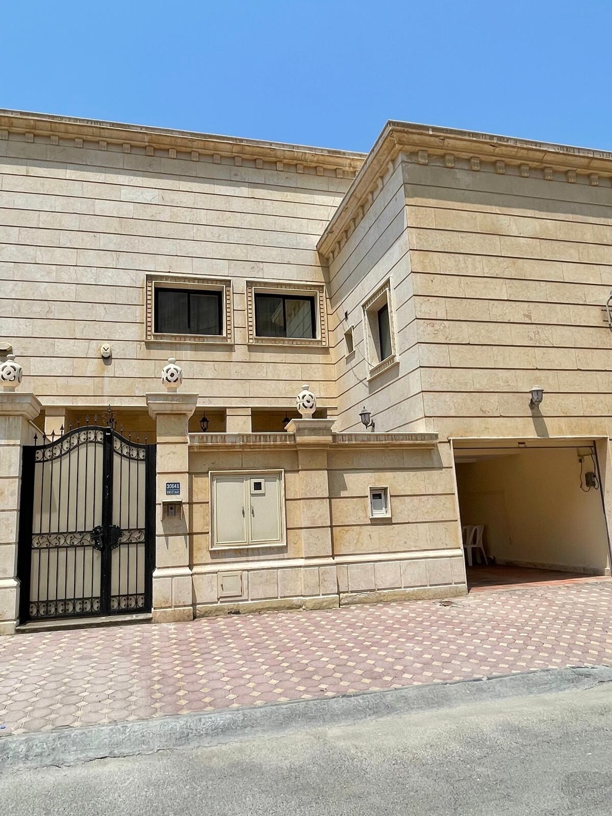 A spacious exterior of a two-story structure is shown, featuring a decorative stone facade and a gated entrance. The driveway leads to a covered parking area, while several windows allow natural light into the building. A clear blue sky is visible above.