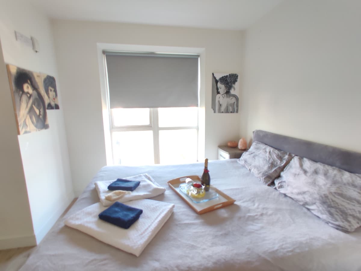 A cozy bedroom is showcased, featuring a spacious bed with soft linens. A tray with a bottle and glasses sits atop the bed, complementing the inviting atmosphere. Natural light filters through a partially closed window with a roller shade, and artistic wall decor adds character.