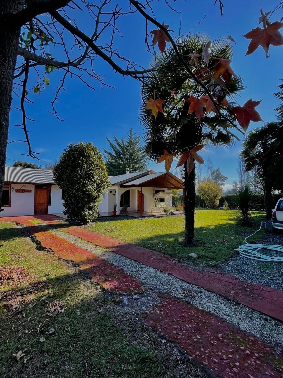 The exterior of a two-bedroom house is showcased, surrounded by well-maintained gardens. A pathway leads to the entrance, framed by trees and ornamental plants. Bright blue skies and sunlight enhance the inviting atmosphere, highlighting the home’s peaceful setting.