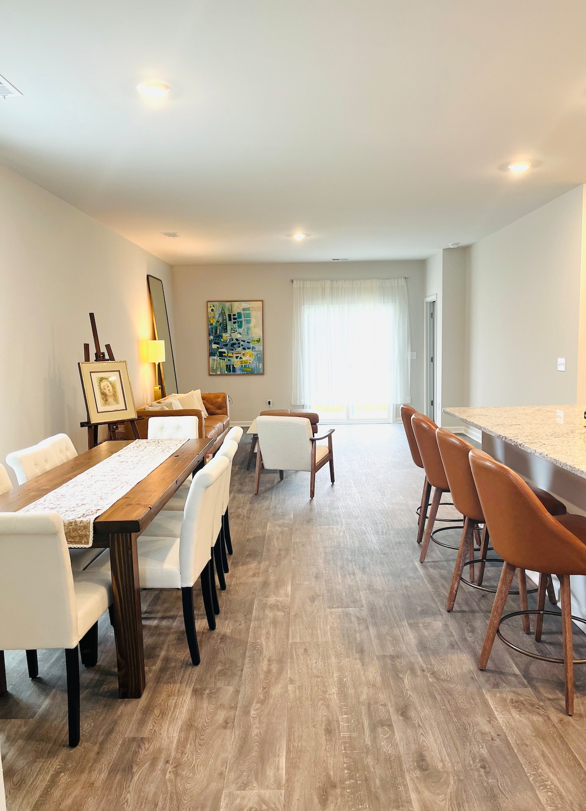 A dining area features a long wooden table accompanied by white upholstered chairs, while additional seating includes modern orange barstools lined up at a granite countertop. Natural light filters through large glass doors, emphasizing the spaciousness of the room and warm neutral tones of the decor.