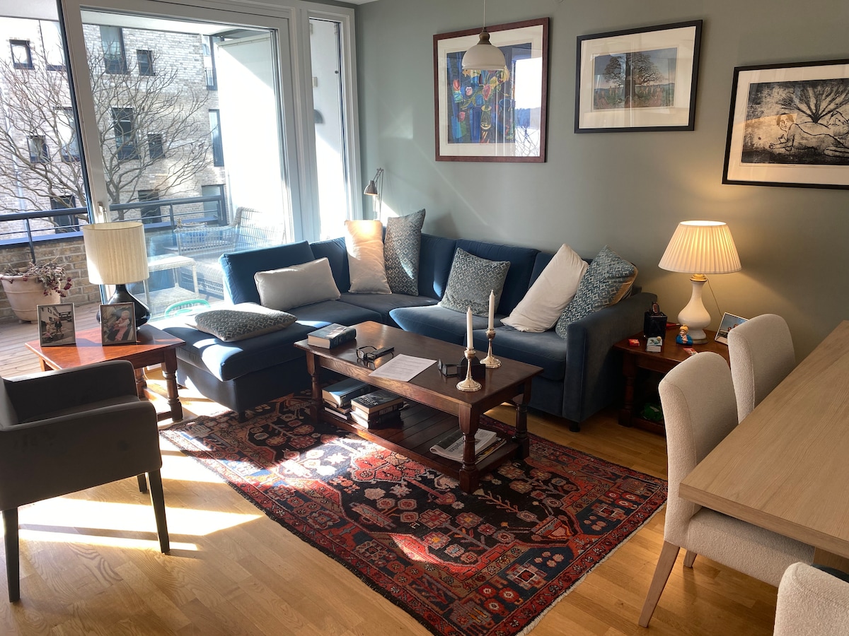 A cozy living room features a modern sectional sofa adorned with decorative pillows. A wooden coffee table holds candles, and soft light emanates from a nearby lamp. An elegant dining table with upholstered chairs sits against a wall, while large windows provide natural light and views.