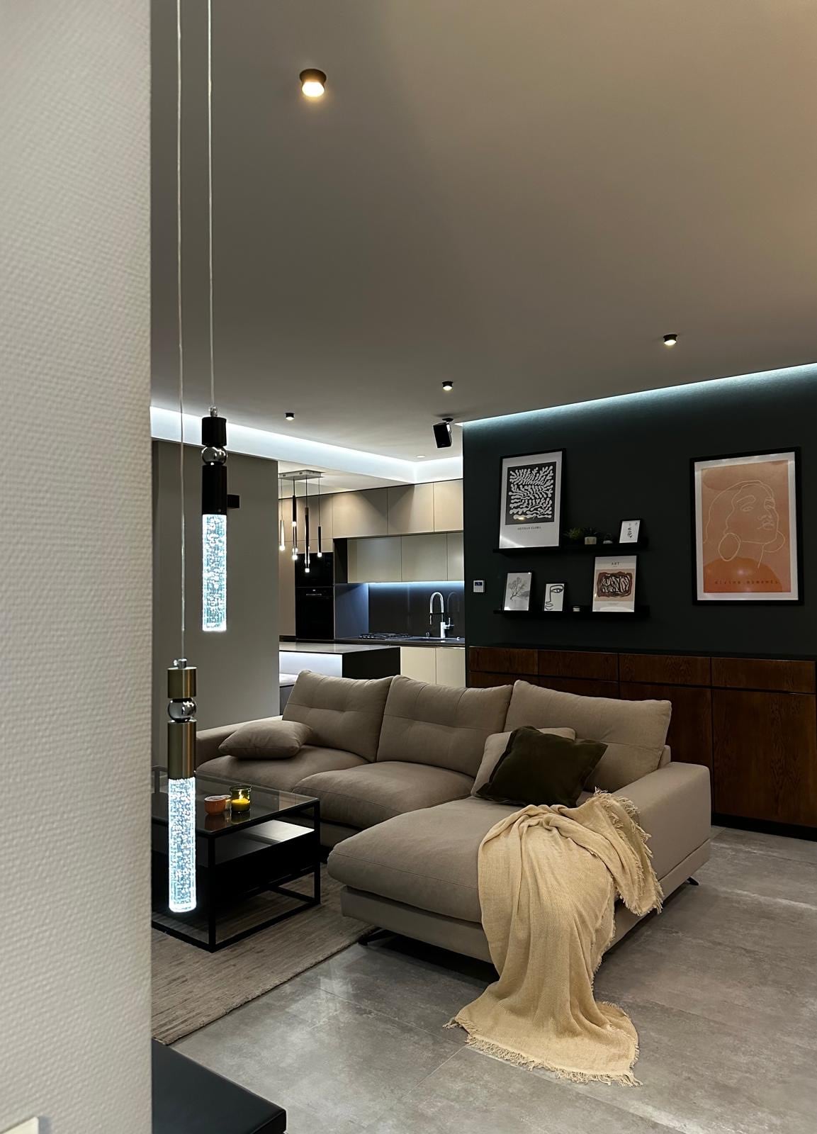 A modern living area features a spacious sectional sofa adorned with a neutral throw blanket. Soft lighting enhances the room, highlighting a low coffee table and a sleek cabinet against the wall. The kitchen is visible in the background, showcasing contemporary cabinetry and appliances.