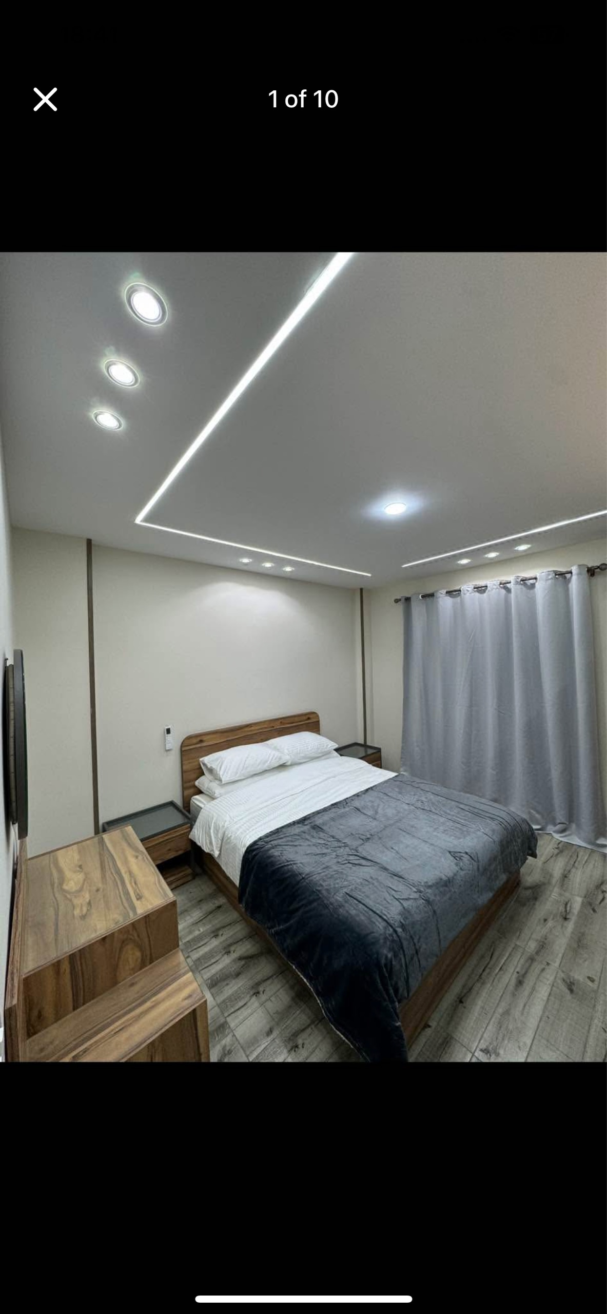 A cozy bedroom features a spacious bed adorned with crisp white linens and a soft gray throw blanket. Soft lighting is provided by recessed lights in the ceiling. A dresser is positioned next to the bed, while sheer curtains frame a window that allows natural light to enter.