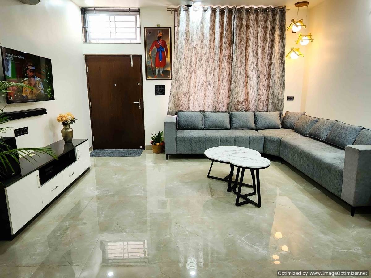 A modern living area features a spacious gray corner sofa, complemented by a circular coffee table. A television is mounted on a sleek white console, while light filters through elegant curtains. Wall art adds a touch of character, enhancing the overall ambiance.