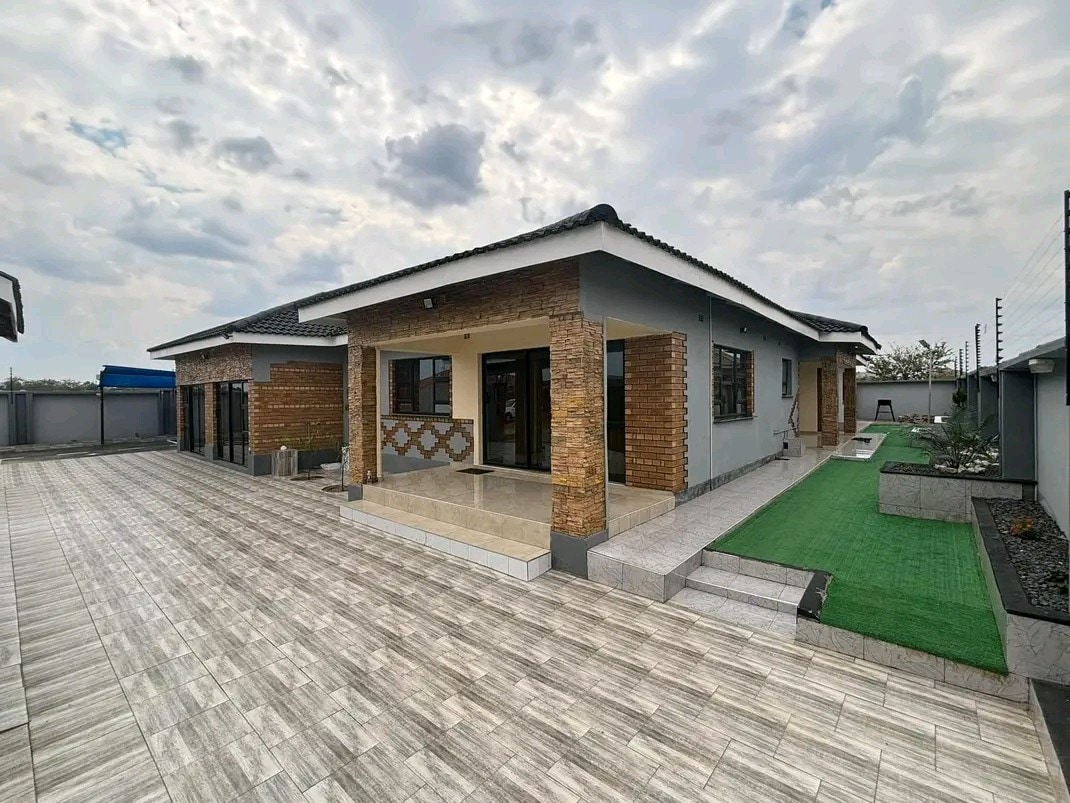 A spacious outdoor area surrounds a single-story home, featuring a combination of textured stone and modern finishes. Paved pathways lead to green lawn spaces, while various textures of the exterior walls create visual interest under a cloudy sky.