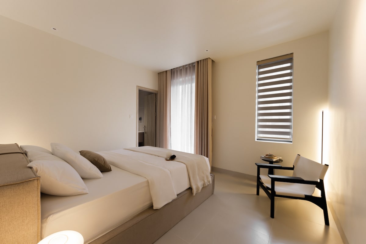 A bright sleeping area is featuring a spacious bed with soft linens and multiple pillows. A chair is positioned nearby, with a small table holding an object. Natural light filters through a window covered by stylish blinds, and light curtains add a soft touch to the room.