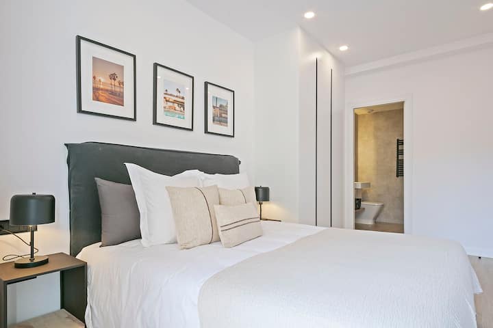 Chumba cha kulala chenye mwanga, cha kisasa chenye kitanda cha watu wawili, mashuka ya kawaida, kichwa cha kitanda kilichofunikwa, meza za kando ya kitanda zilizo na taa na mchoro ulio juu. Kuta nyeupe, makabati yaliyojengwa ndani na bafu la chumbani linaloonekana kupitia mlango.