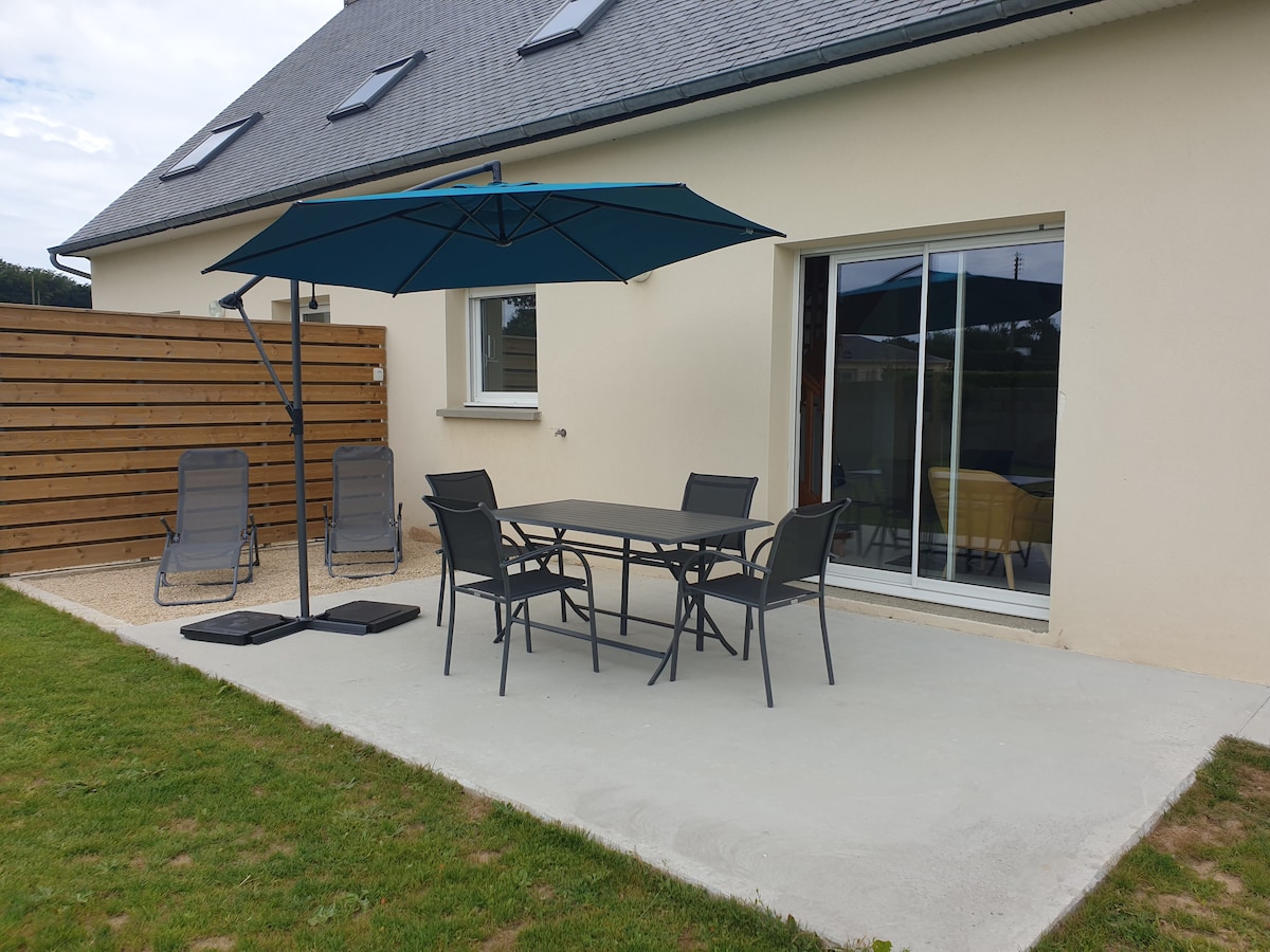 An outdoor patio area features a table with four chairs, accompanied by two lounge chairs. A large umbrella provides shade to the table. The space is bordered by a wooden fence and a well-kept lawn, with access to the interior through sliding glass doors.