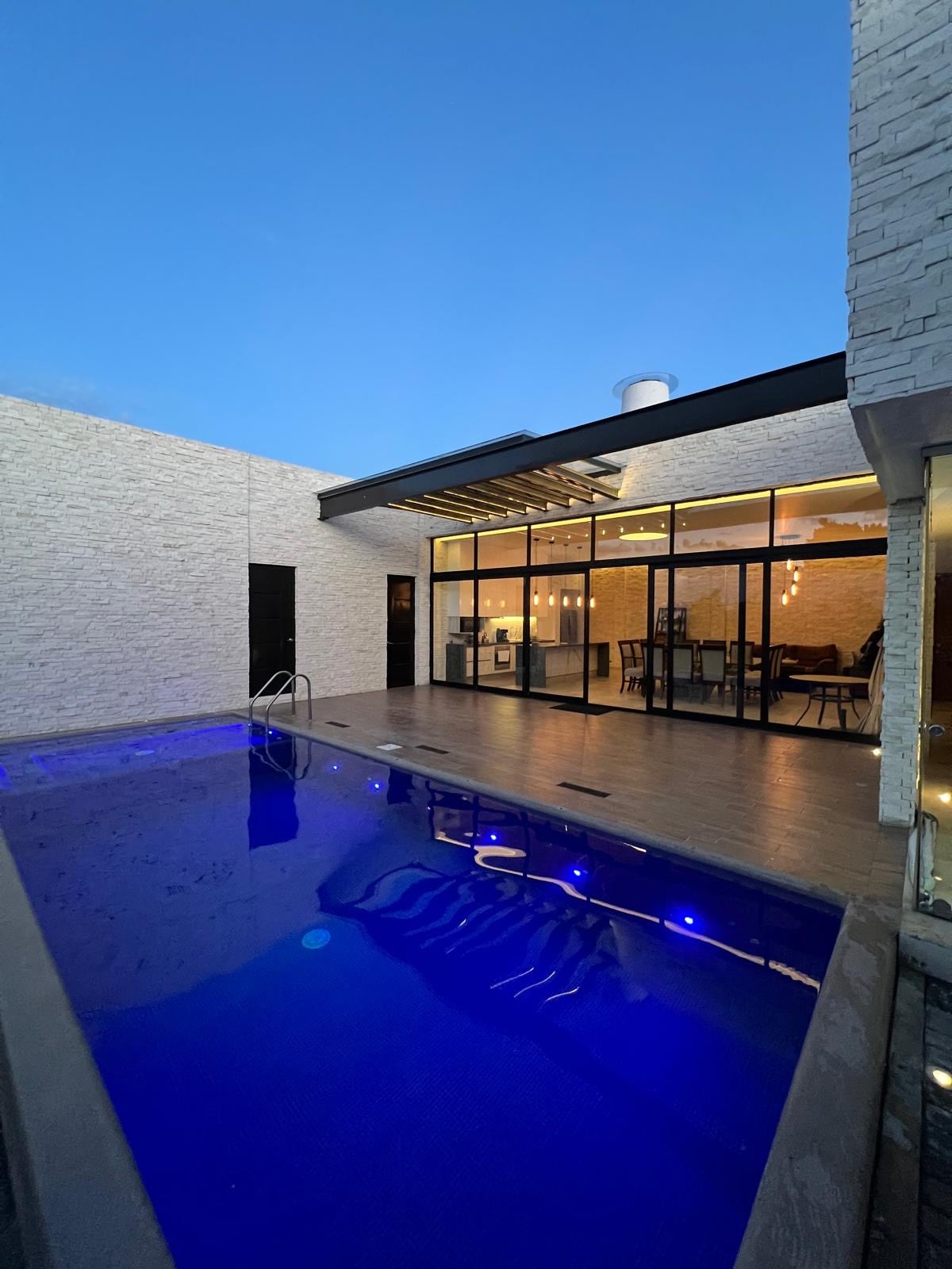 A contemporary outdoor space features a luminous pool complemented by soft blue lighting. Large glass doors provide a view into the elegantly designed interior, while a sleek roof structure offers shade. The surrounding area is finished with light-colored stone walls, contributing to an open ambiance.