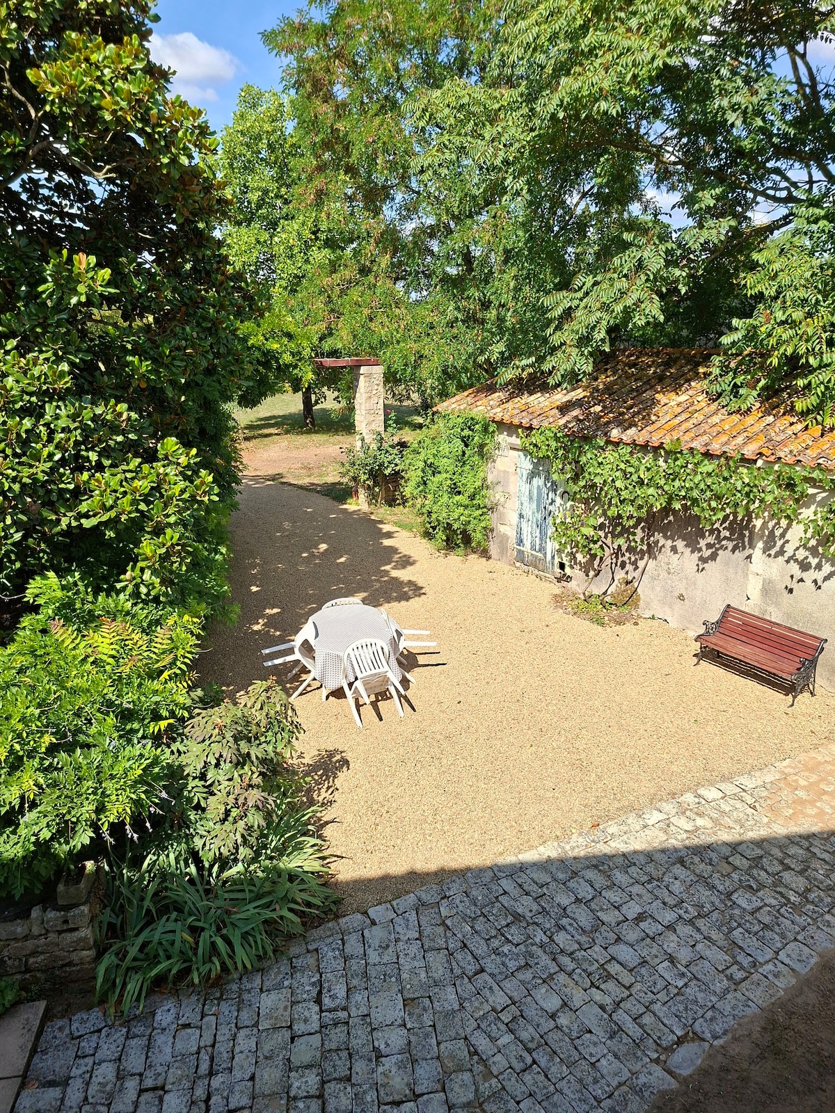 A peaceful outdoor area is featured, surrounded by lush greenery and a gravel pathway. A circular table with white chairs is positioned in the center, complemented by a wooden bench nearby. An old stone building with blue doors is visible, along with trees providing shade.