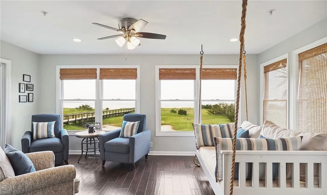 A bright and airy sitting area features large windows with bamboo shades, showcasing views of the outdoor landscape. Two comfortable chairs and a sofa are arranged for relaxation. A swing bed with decorative pillows offers a unique touch, inviting moments of leisure.