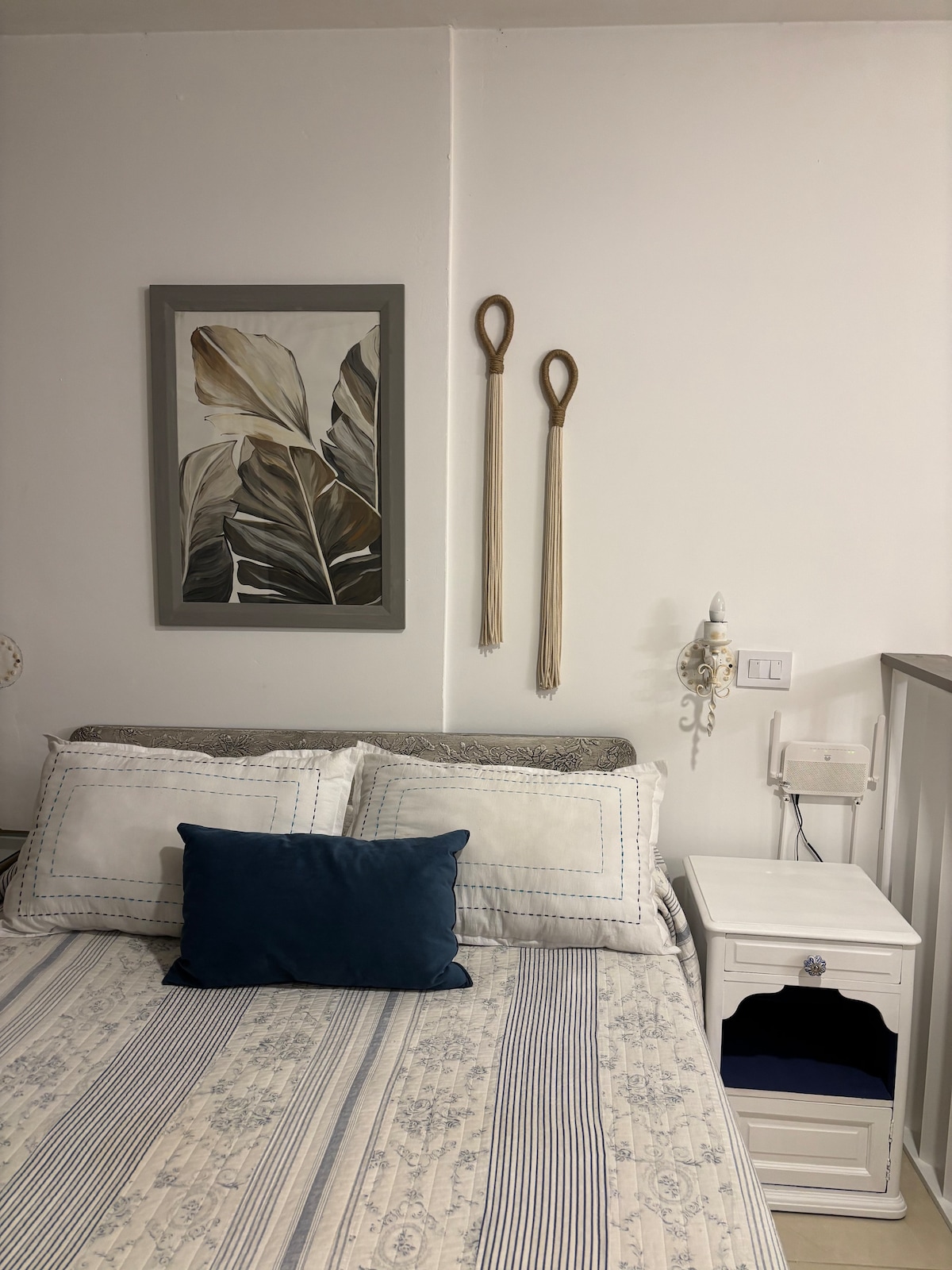 A comfortable sleeping area showcases a spacious bed adorned with decorative pillows. A wall-mounted artwork featuring leaves adds a natural touch. A small nightstand is positioned beside the bed, with subtle lighting fixtures enhancing the setup.