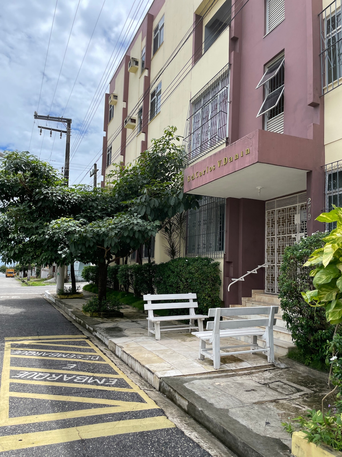 A two-story building is featured, with a welcoming entrance adorned by a decorative wrought iron gate. Two white benches are placed along a stone walkway, surrounded by greenery. The scene is complemented by a clear sky and utility poles lining the street.
