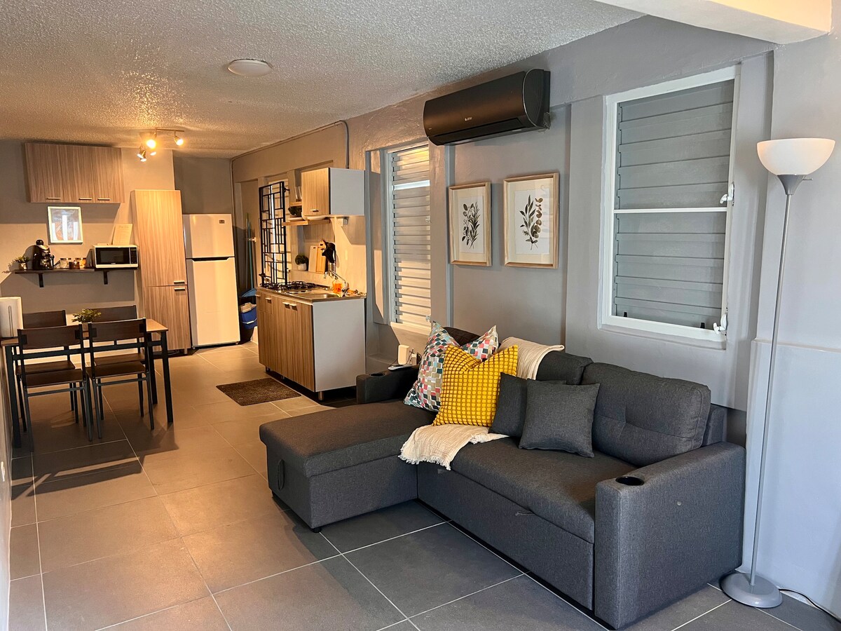 A modern living space is showcased, featuring a spacious gray sectional sofa adorned with decorative pillows. A dining area with a table for four is visible, along with a fully equipped kitchen. Light streams in through the windows, enhancing the neutral color scheme of the room.
