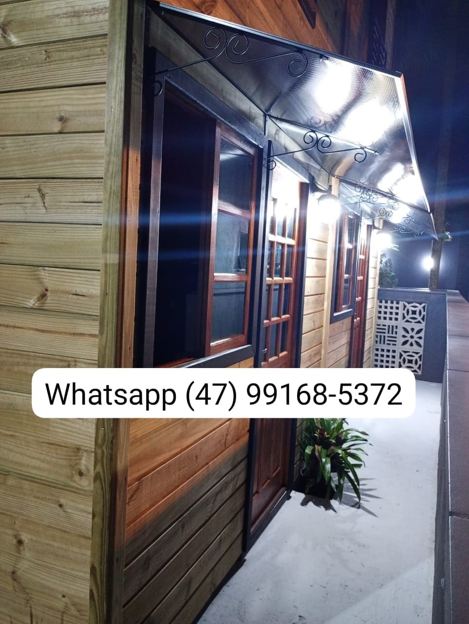 The exterior of the two-story cabin is shown, featuring warm wooden siding and a covered entrance. Soft lighting illuminates the porch area, highlighting a small potted plant near the entrance. The presence of a contact number is noted on the image.