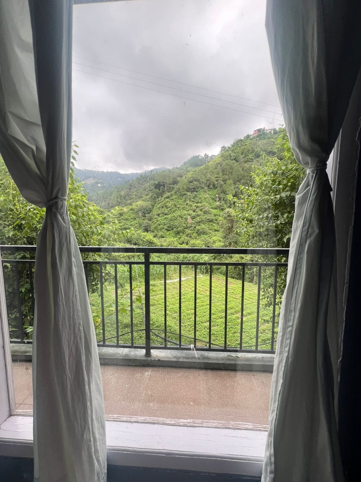 A wide window framed by sheer curtains opens to a lush, green landscape. Vibrant hills and a distant row of trees are visible, providing a calming view of nature. The balcony railing complements the serene outdoor scene.