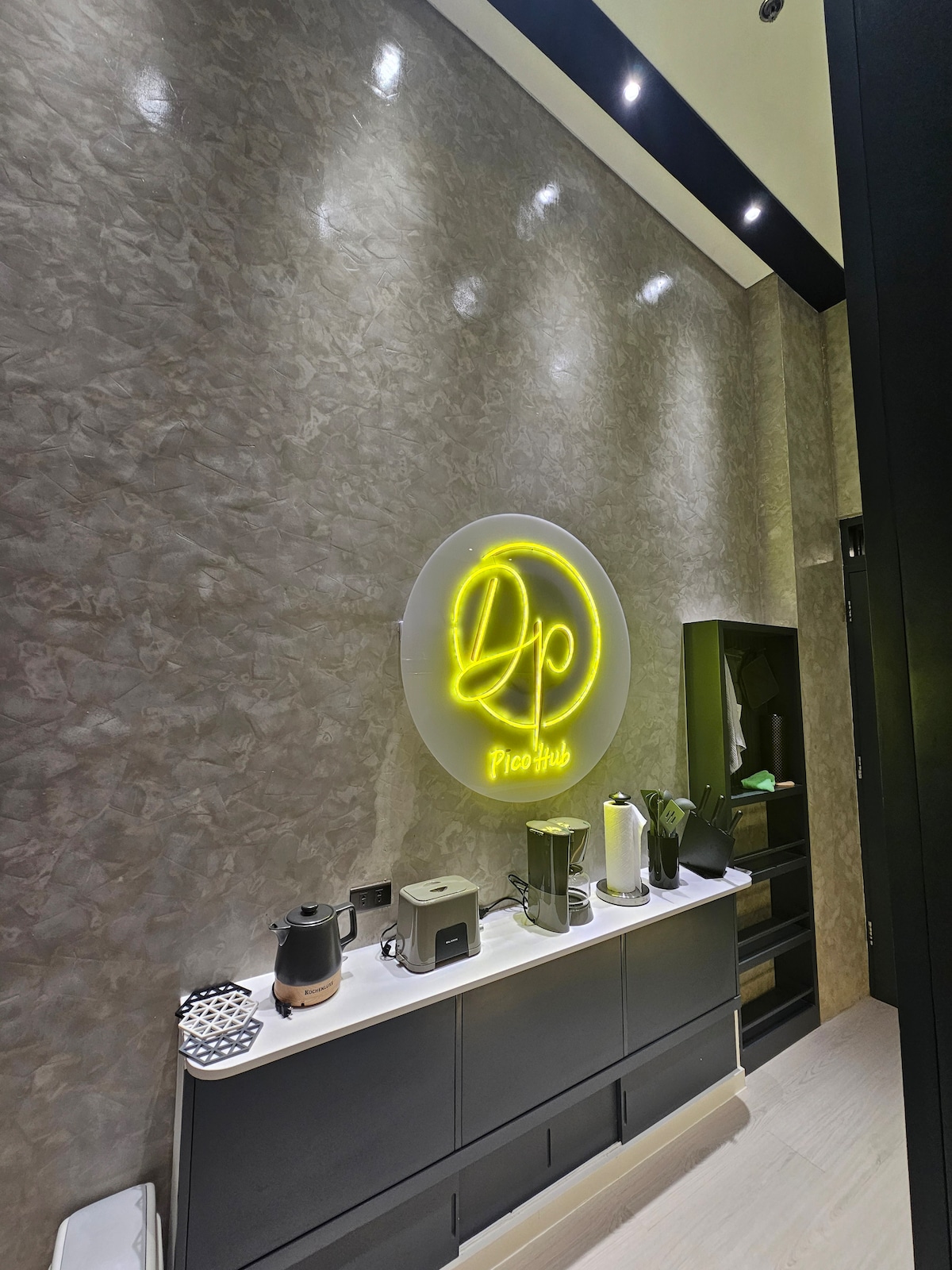 A modern kitchenette is highlighted by a glowing neon sign, with sleek cabinetry and a countertop displaying a coffee maker and several kitchen essentials. Subtle lighting accentuates the textured walls, while a dark storage area is visible in the background.