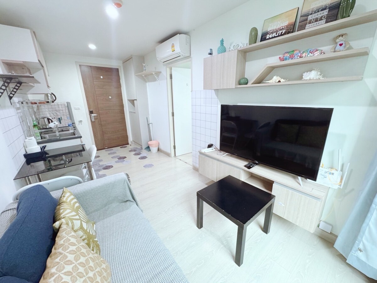 A modern living area features a comfortable couch adorned with decorative pillows and a sleek coffee table. A wall-mounted television is prominently displayed on a light-colored media console. The space is illuminated by soft lighting and includes wood accents for a cozy touch.