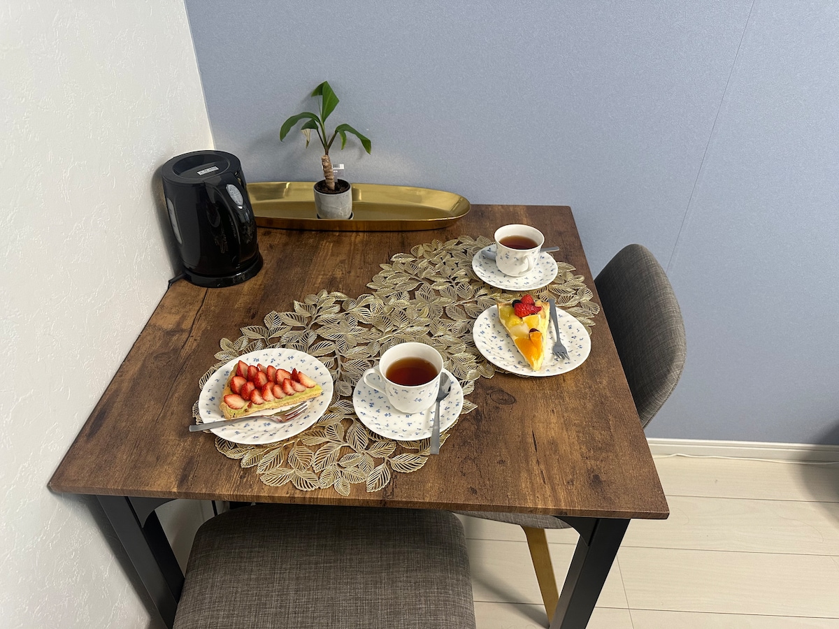 A wooden dining table is set for two with plates of fruit and desserts. A teapot accompanies cups of tea, and a small potted plant is placed on the table. A sleek black kettle is positioned nearby, enhancing the inviting dining space.