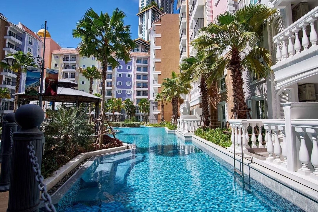 An inviting lagoon-style swimming pool is surrounded by tropical palm trees and colorful buildings. The clear blue water reflects the serene environment, complementing the vibrant landscaping and modern architecture of the surrounding area.