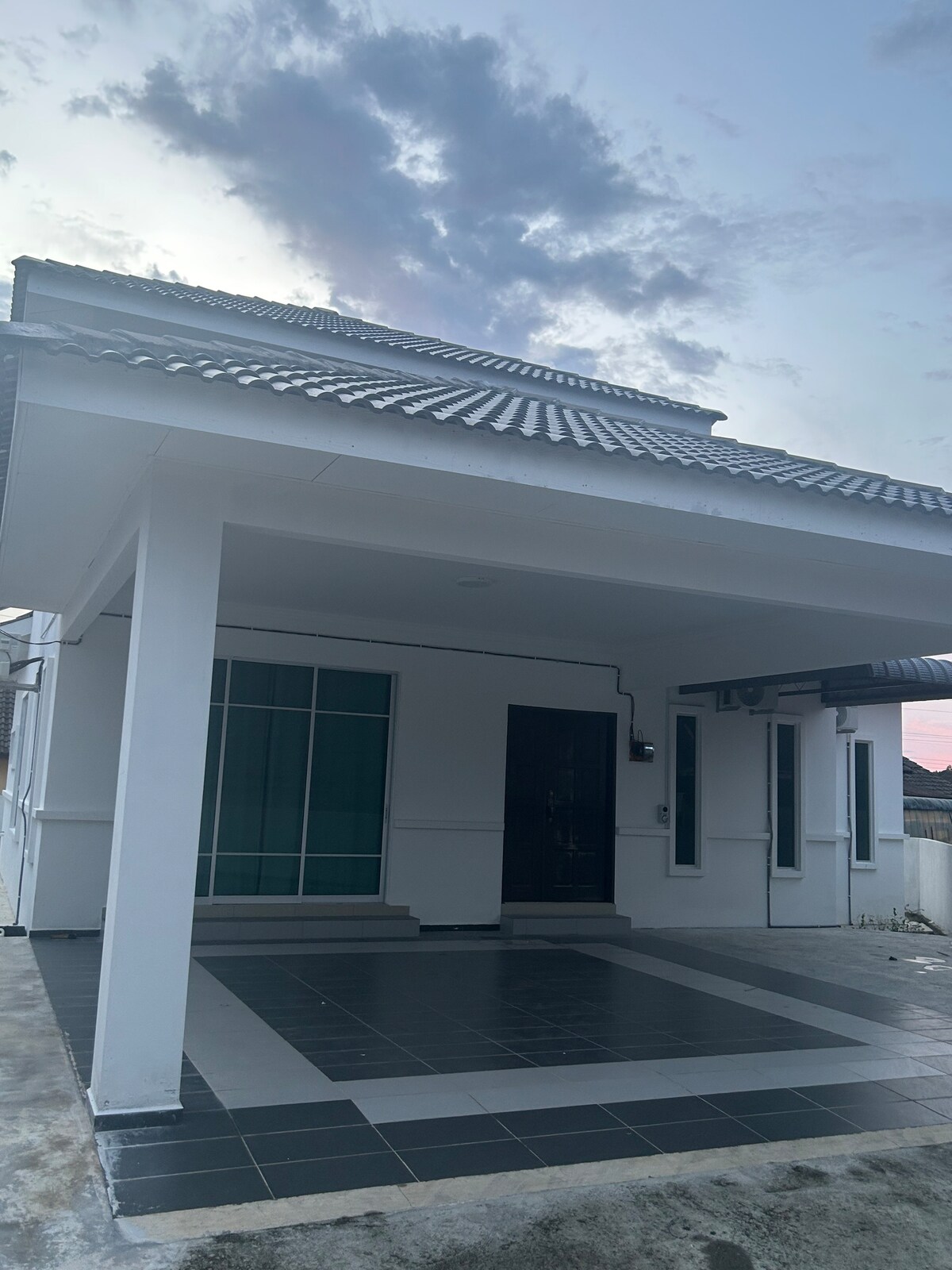 A modern house is presented with a sleek exterior of white and dark grey tiles. The entrance features a spacious porch with large windows allowing natural light to fill the front room. A tiled pathway leads to the main door, framed by a textured roof.