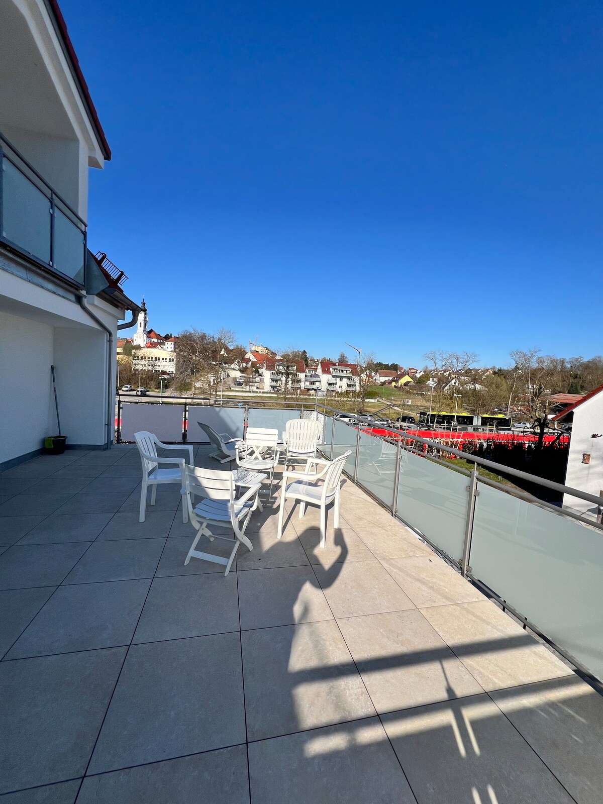 A spacious terrace is shown, featuring several white outdoor chairs arranged around a table. A glass railing offers a clear view of the surrounding landscape, which includes buildings and greenery under a clear blue sky. Sunlight is present, creating a bright and open atmosphere.