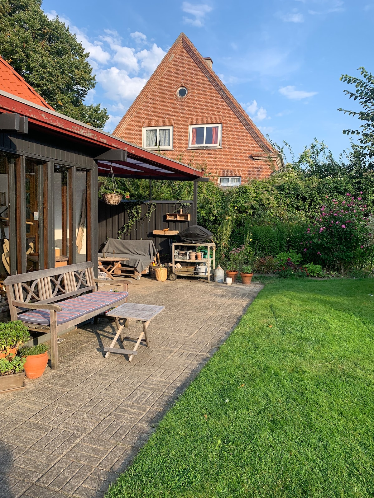 A patio area is visible with wooden furniture arranged on tiled flooring. Green grass serves as a natural border, with various potted plants positioned around. A brick house with multiple windows is situated in the background, framed by trees and shrubbery.