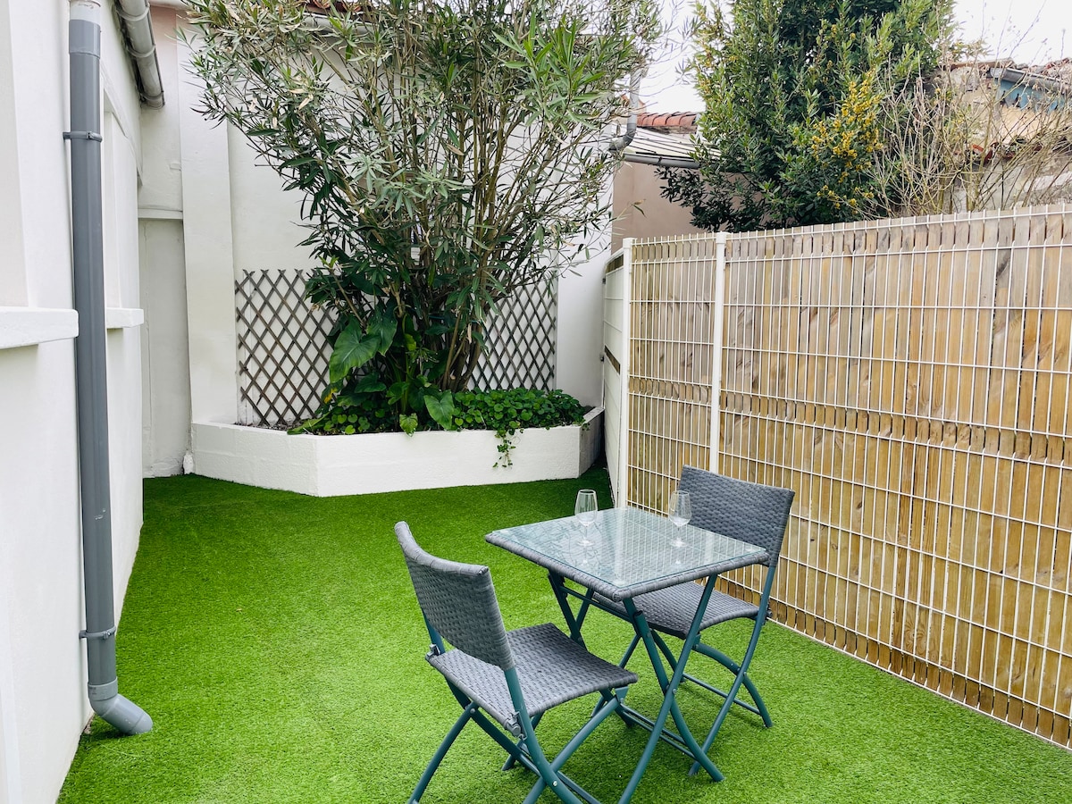 A private outdoor courtyard is depicted, featuring a small glass table surrounded by two folding chairs. Lush greenery, including an olive tree, provides a natural backdrop. The space is framed by a wooden fence and artificial grass, creating a peaceful and inviting atmosphere.