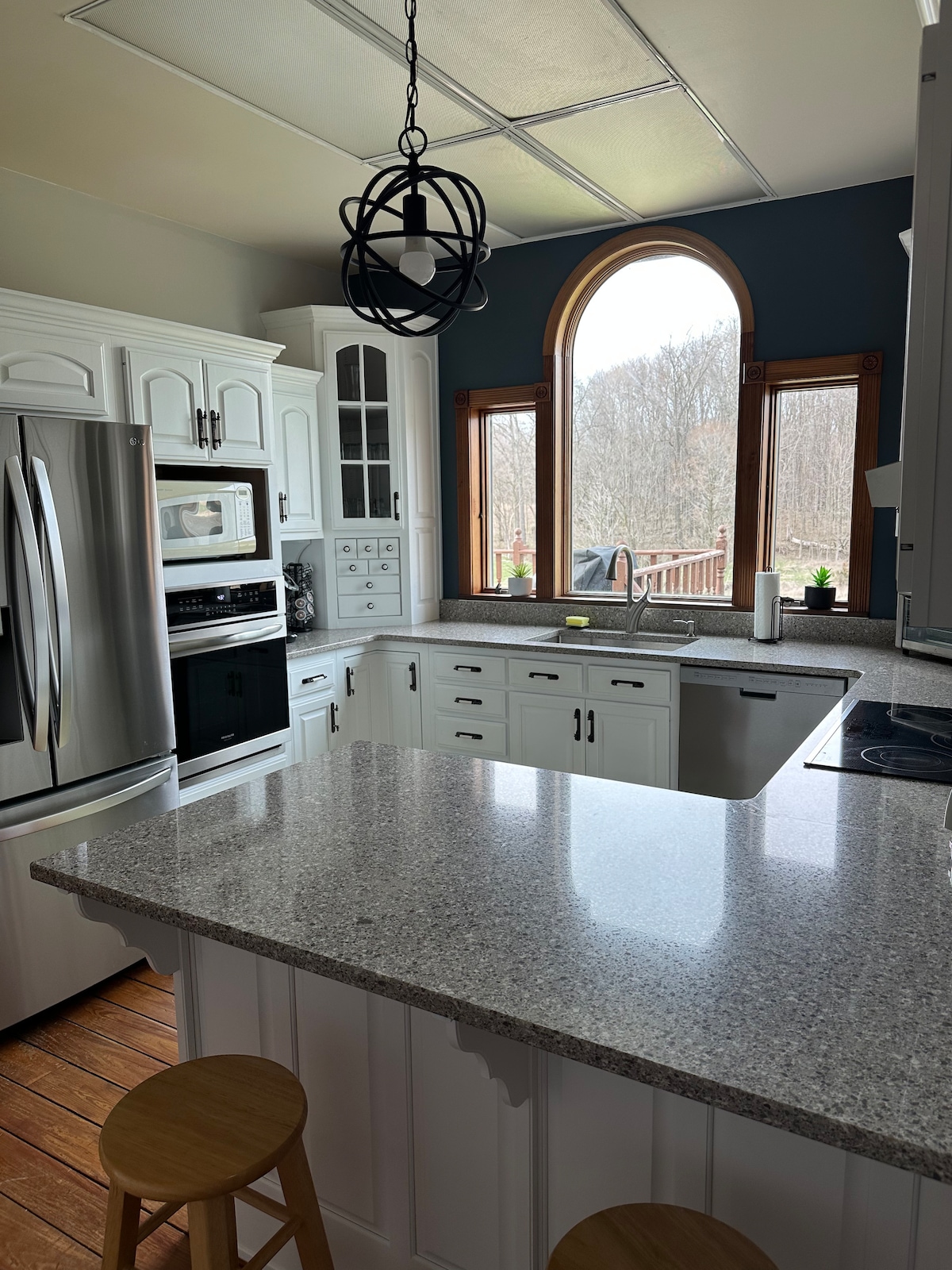 A bright kitchen features white cabinetry, stainless steel appliances, and a spacious granite countertop. A large window with an arched design allows natural light to filter in, while two wooden stools are positioned at the countertop, providing a casual seating area.
