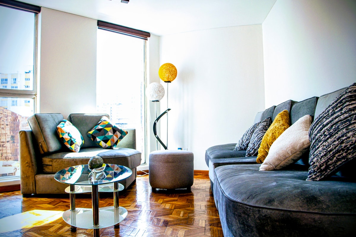 A spacious living area features large windows allowing natural light to fill the space. Comfortable seating options are arranged around a round glass coffee table, with decorative pillows adding subtle color. A floor lamp and a sculptural light source stand to enhance the ambiance.