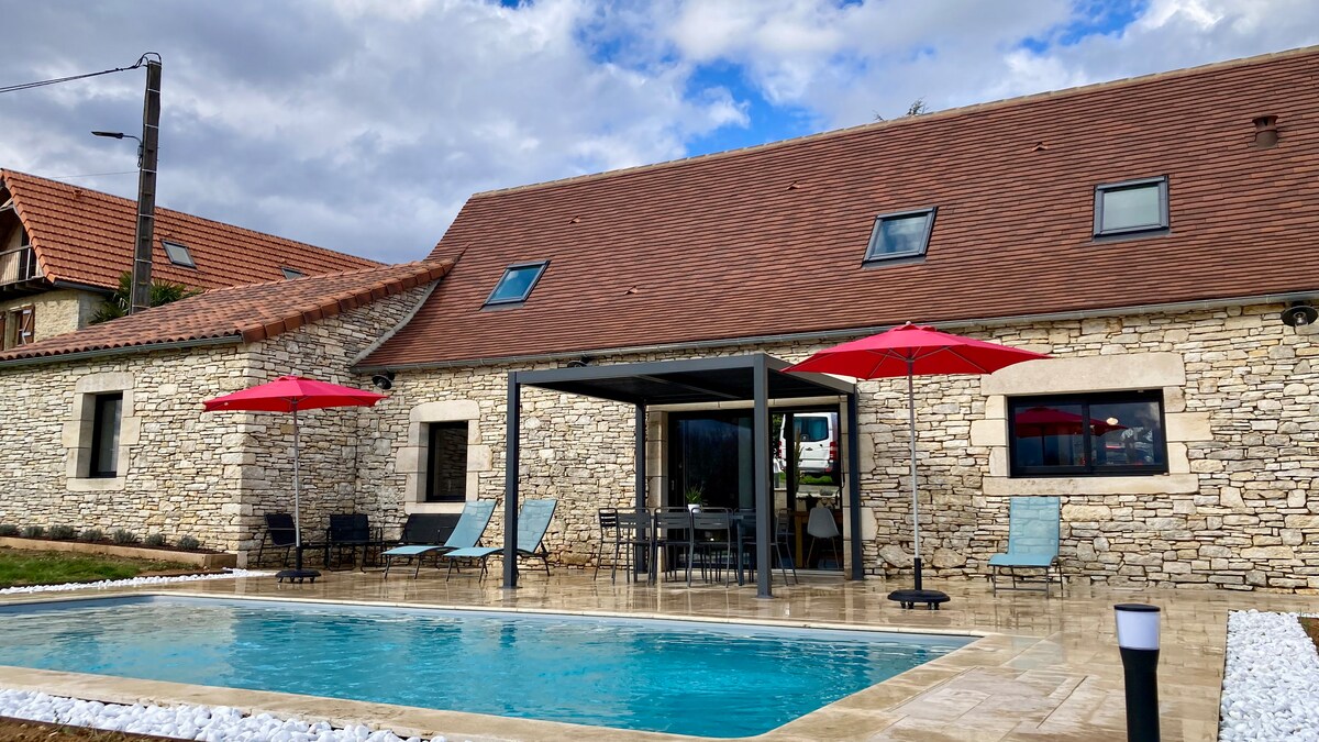 The outdoor area features a rectangular private pool surrounded by a stone patio. Two red umbrellas provide shade over the seating area with lounge chairs. The house, constructed of light stone, displays multiple windows and a well-maintained lawn in the background.
