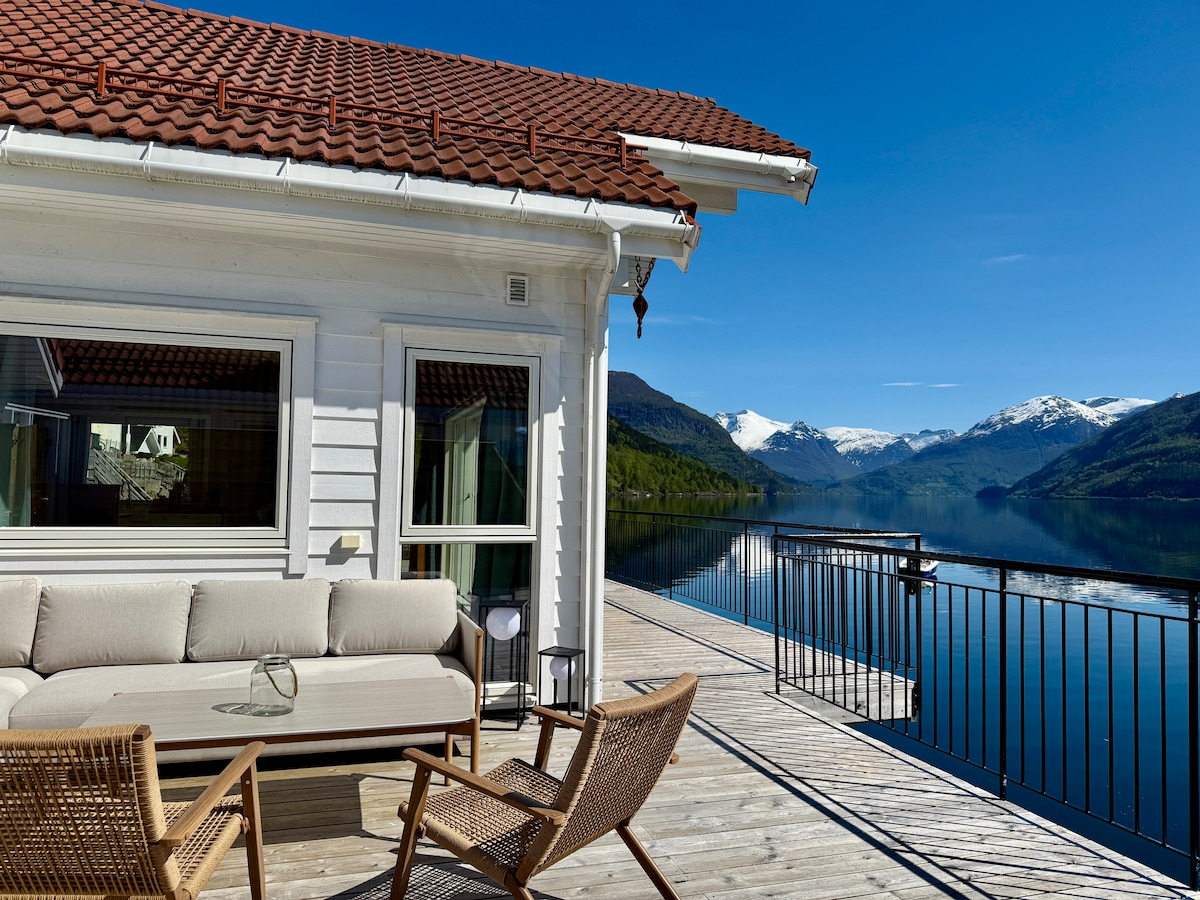 A spacious terrace overlooks the water, featuring cozy seating arrangements with a comfortable sofa and armchairs. The serene landscape includes a panoramic view of mountains and a clear sky, enhancing the tranquil setting by the fjord.