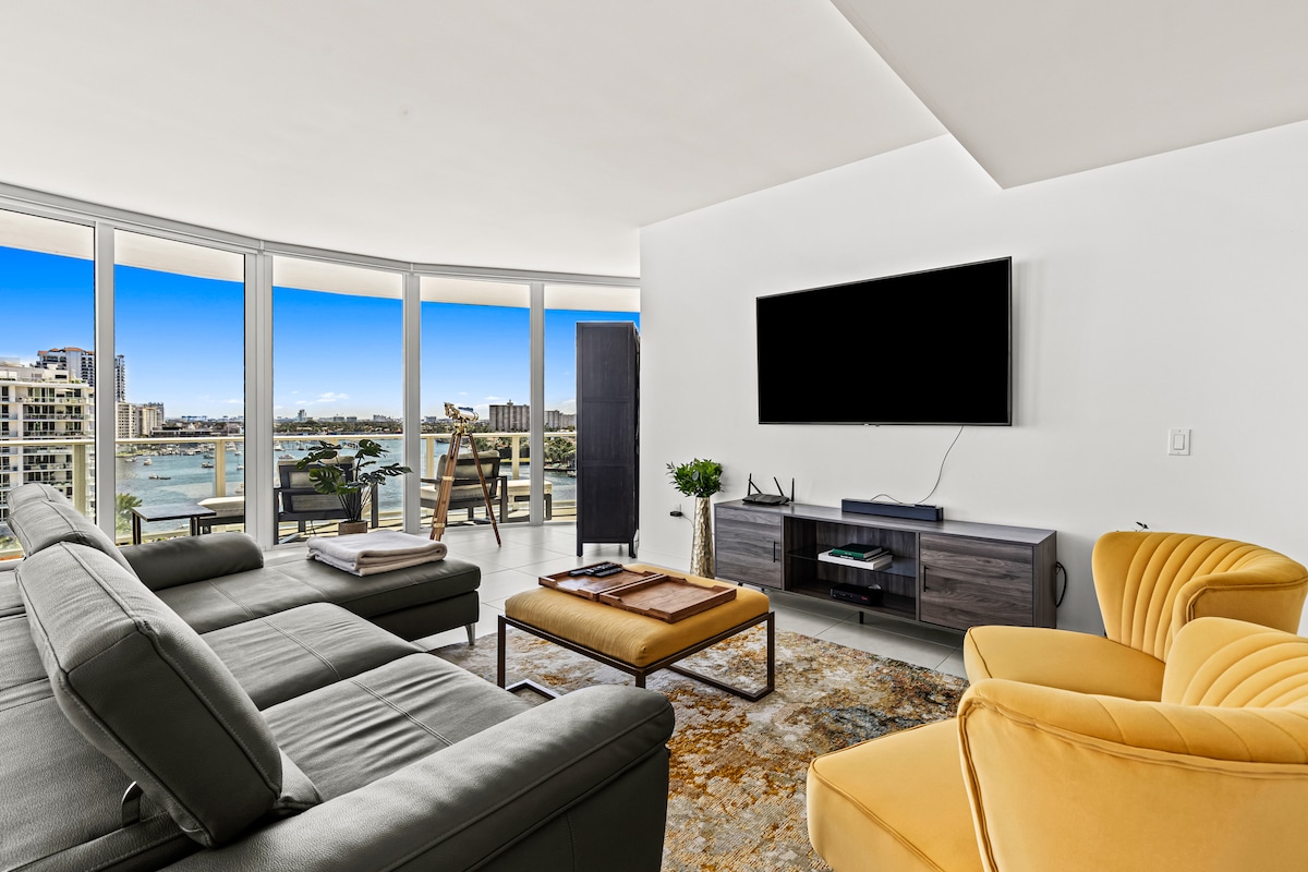A modern living area features a sleek grey sectional sofa adjacent to a stylish yellow accent chair. A coffee table sits in the center, complemented by a decorative area rug. A large flat-screen TV is mounted on the wall, with expansive windows showcasing scenic views.