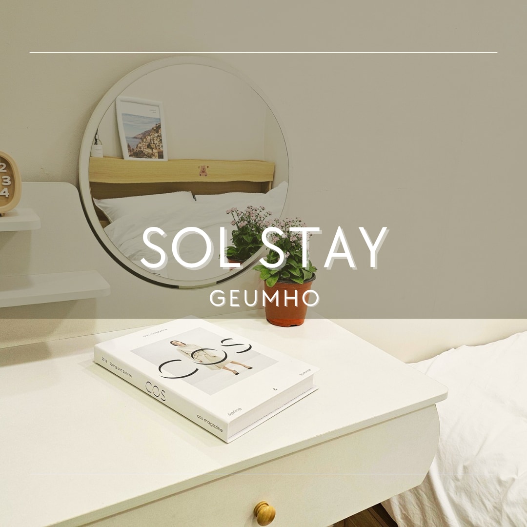 A neatly arranged bedside table is featured, displaying a book atop a white surface. A small potted plant adds a touch of greenery. A round mirror reflects the light-colored decor, contributing to a minimalist and calming ambiance.
